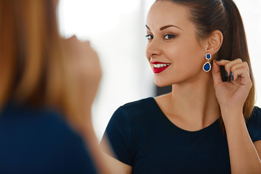 Fashion Woman Portrait. Beautiful Elegant Successful Female With Evening Make-up Wearing Luxurious Blue Dress Smiling In The Mirror. Jewelry And Beauty. Wellbeing, Luxury Lifestyle.