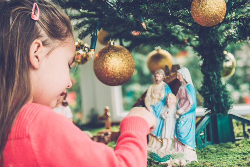 Cute girl of four in pink playing with figurines at Nativity scene, balcony, Primorska, Slovenia, Europe. Holy family figurines, Christmas Tree with golden baubles on the moss. No snow, mild climate. Nikon D800, full frame, XXXL.