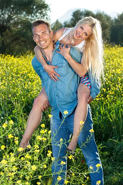 Handsome young man with his pretty blond girlfriend having a relaxing day in the countryside giving her a piggy back ride through the yellow flowers of a rapeseed field.