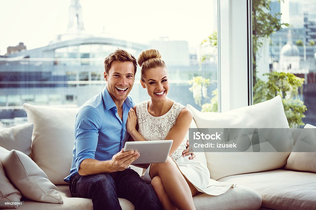 Happy couple with a digital tablet Young couple sitting on sofa in an apartment, holding a digital tablet and laughing at the camera. Couple - Relationship Stock Photo