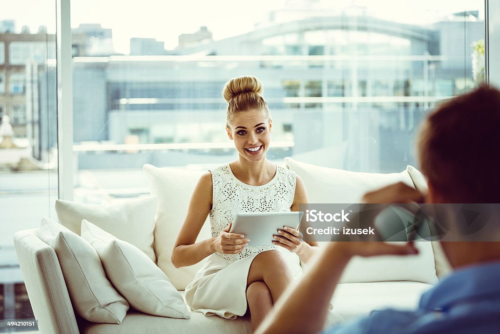 Woman with digital tablet Focus on young woman sitting on sofa at home and using a digital tablet with the back side view of a men talking on phone on the foreground. 25-29 Years Stock Photo