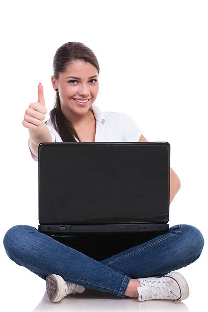 casual young woman sitting with legs crossed and showing thumb up while holding laptop. isolated on white background