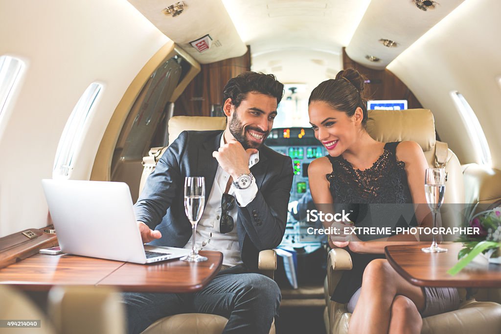 Business couple enjoying in private aeroplane Young business couple in private aeroplane during the flight having a champagne and smiling while looking at computer screen. Cockpit is in the background. Couple - Relationship Stock Photo