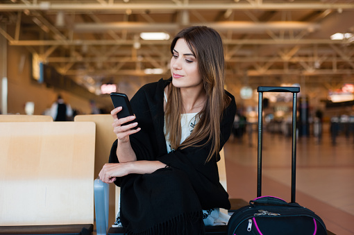 Airport Young female passenger on smart phone at gate waiting in terminal while waiting for her flight. Air travel concept with young casual woman sitting with hand luggage suitcase.