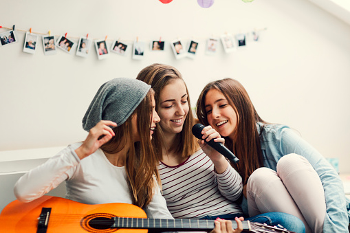 Teenage Girls Having fun together at slumber party. Sitting on couch and playing guitar and singing karaoke. Shot with Canon EOS 5Ds 50mp. Selective focus to girl with microphone.