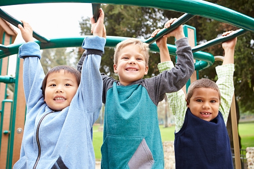 Three Young Boys On Climbing Frame In Playground Smiling To Camera
