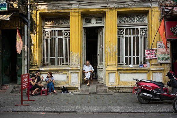 Life in old town Hanoi, Vietnam - May 3, 2014: Old man reads book in front of ancient house, with some teens drink tea nearby in Hang Than str, Hanoi, Vietnam vietnamese old man portrait stock pictures, royalty-free photos & images