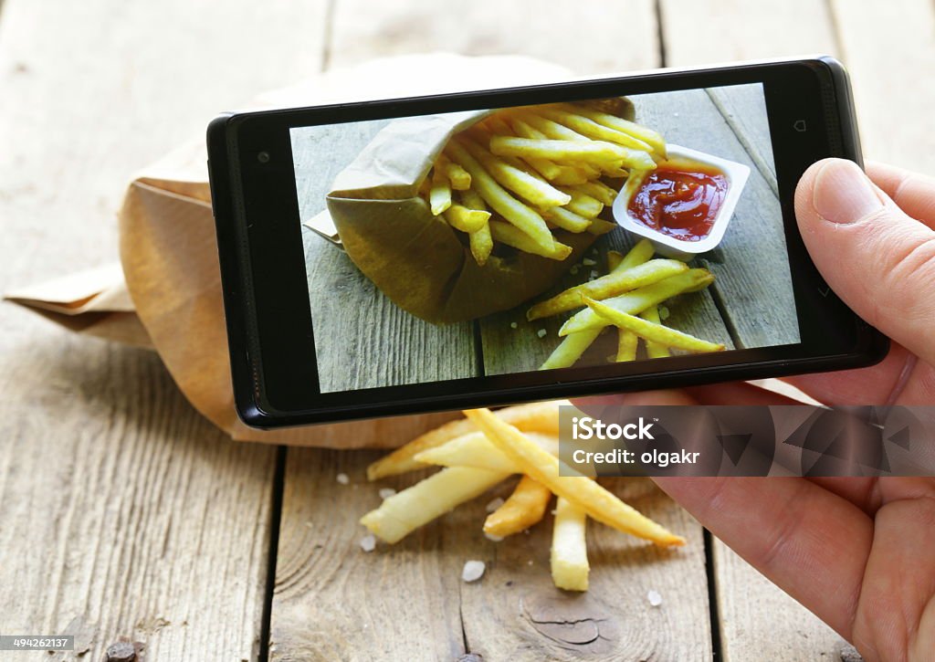 French fries on mobile phone screen A smartphone being used to snap a photo of some French fries is pictured.  The French fries are in a cardboard container, and they have a disposable container of ketchup next to them.  They are on a wood table.  The smartphone obstructs the view of the French fries. Auto Post Production Filter Stock Photo