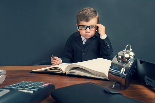 Young boy dressed in a suit working at a large desk. The desk has a calculator, Filofax, phone and clock. He is very busy and there is a lot of work on his desk. He is wearing glasses and looks like an accountant.