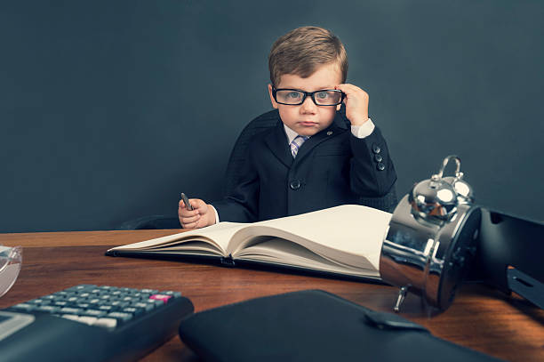 Young boy dressed in suit working at desk. Young boy dressed in a suit working at a large desk. The desk has a calculator, Filofax, phone and clock. He is very busy and there is a lot of work on his desk. He is wearing glasses and looks like an accountant. bossy photos stock pictures, royalty-free photos & images