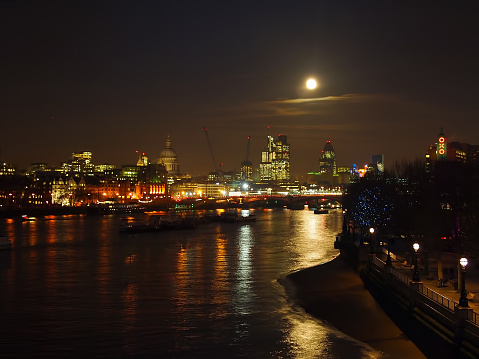 Long exposure across the edge of Waterloo Bridge with a view of St Paul's Cathedral, the Gherkin building and South Bank with a full moon in the sky.