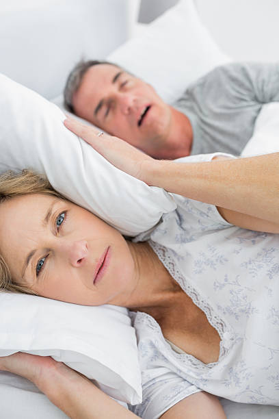 Tired wife blocking her ears from noise of husband snoring Tired wife blocking her ears from noise of husband snoring looking at camera in bedroom at home camera covers pictures stock pictures, royalty-free photos & images