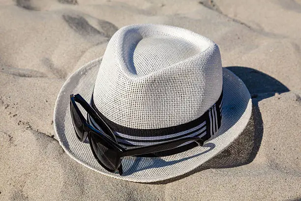 White straw fedora hat on sandy beach with black rimmed polarized sunglasses.