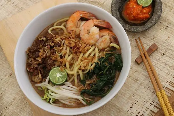 A Peranakan dish from Betawi ethnic in Jakarta, noodle soup with water spinach as the main vegetable, with sweet soy braised beef, shrimps, beansprouts in hot beef broth. Served fancy style in plain white ceramic bowl, with chili paste and kaffir lime aside. A pair of wooden chopsticks on chopstick rest is placed on the right hand side of the bowl. The bowl is placed on an elevated wooden board. A woven straw mat underline the dishes. The image was for overhead view; indoor near window and used sunlight as the main lighting source. 