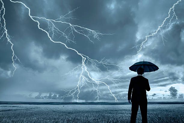 Man in black suit with a black umbrella in a storm A rear view of a businessman in a suit, with an upheld umbrella, standing in a large field during a thunderstorm. Lightning is seen descending from a gray and cloudy sky. emergency shelter photos stock pictures, royalty-free photos & images