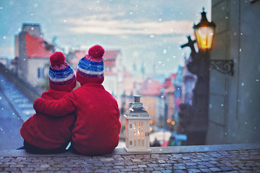 Two cute kids, boys, standing on stairs, holding a lantern, view of Prague behind them, snowy evening