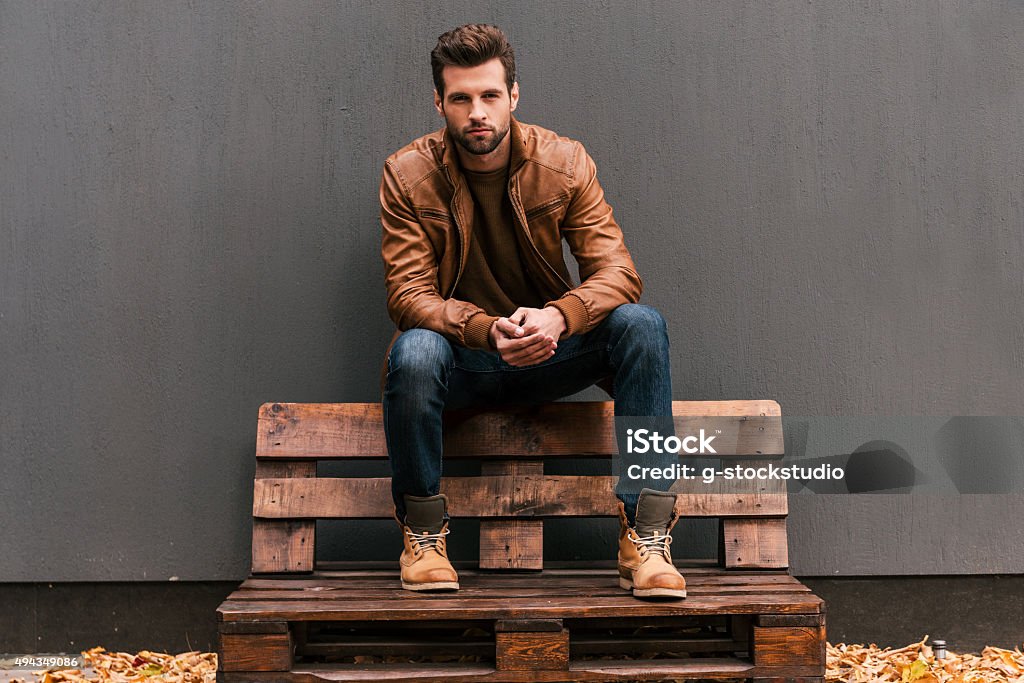 Confident and handsome. Handsome young man sitting on the wooden pallet and looking at camera with grey wall in the background and orange fallen leaves on the floor Men Stock Photo