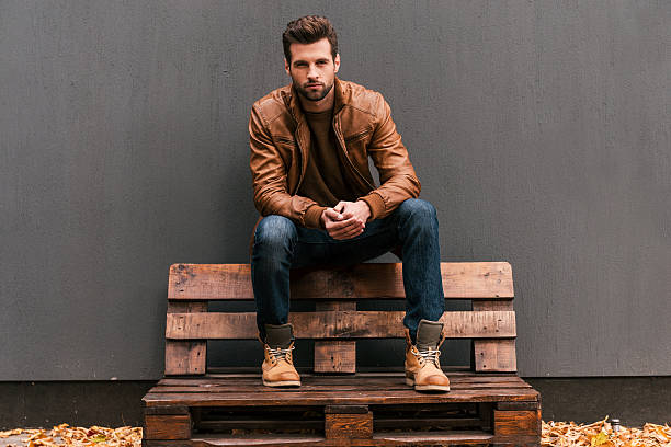 Confident and handsome. Handsome young man sitting on the wooden pallet and looking at camera with grey wall in the background and orange fallen leaves on the floor handsome people stock pictures, royalty-free photos & images
