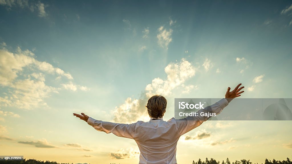 Successful man  standing  with his arms spread towards majestic sky Successful man in elegant white shirt standing with his back to the camera with his arm spread widely towards beautiful majestic evening sky as he celebrates his success and prosperity. Abundance Stock Photo