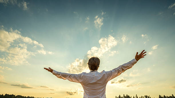 Successful man  standing  with his arms spread towards majestic sky Successful man in elegant white shirt standing with his back to the camera with his arm spread widely towards beautiful majestic evening sky as he celebrates his success and prosperity. prosperity stock pictures, royalty-free photos & images