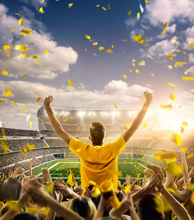 A long-range shot of a stadium field, floodlights and seating. A green field, with painted white lines, is visible in the foreground. On the foreground a group of fans is celebrating. One man stands with his hands up to the sky. People are dressed in yellow colors. In the background are diffuse out-of-focus stadium seats. Large, bright floodlights are in the center part of the image.