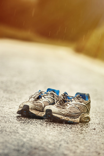 Worn-out mens running shoes standing on a desolate country road in a rainy day. Sports, active lifestyle, running, marathon, individual sports concept.