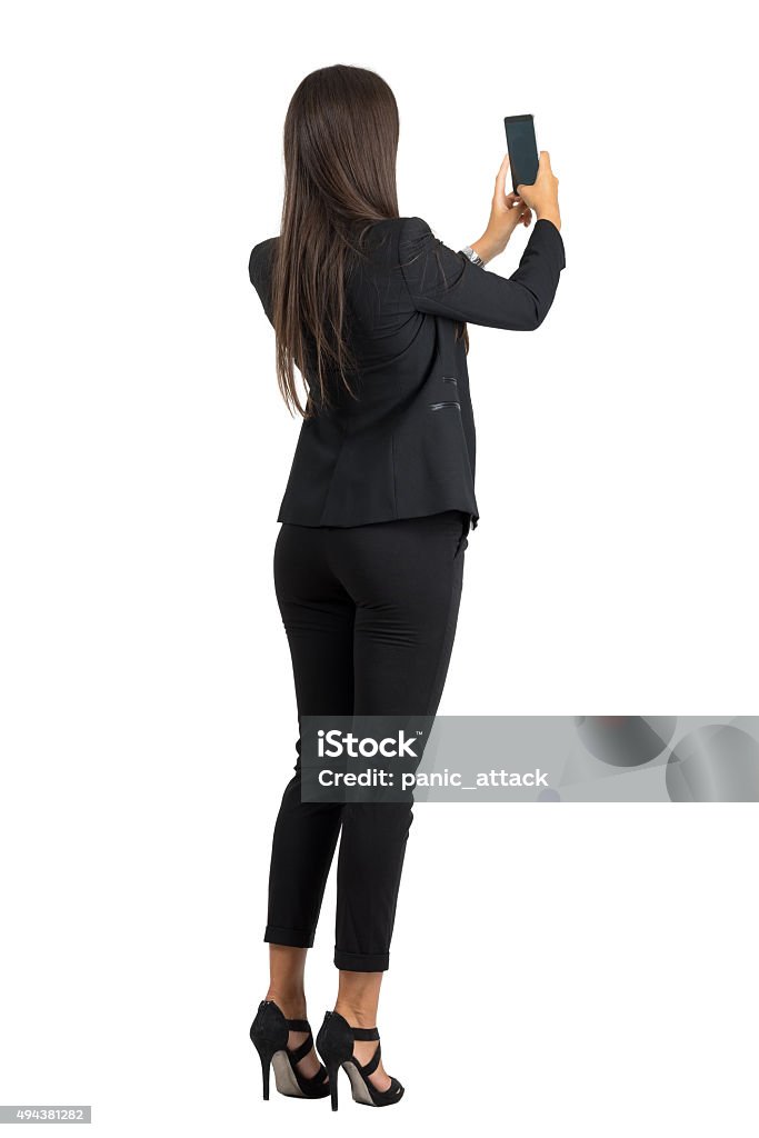 Rear view of woman in suit taking photo with cellphone Rear view of long hair corporate woman in suit taking photo with mobile phone. Full body length portrait isolated over white studio background. Women Stock Photo