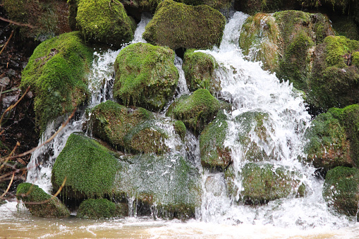 Photo showing a splashing waterfall with moss covered rocks, where the water is cascading into the adjacent river.