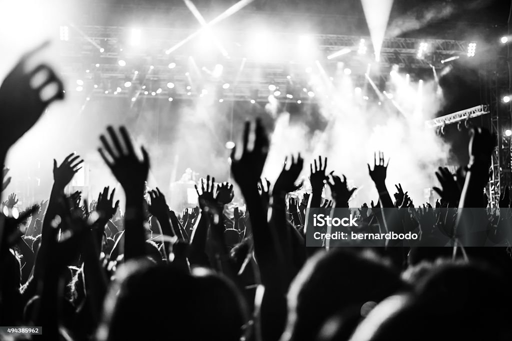 Put your hands up in the air! Black and white photo of audience with hands raised at a music festival and lights streaming down from above the stage. Soft focus, blurred movement. Performance Stock Photo