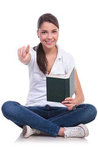 casual young woman sitting with legs crossed and holding a book while pointing and looking at the camera with a smile. isolated on white background