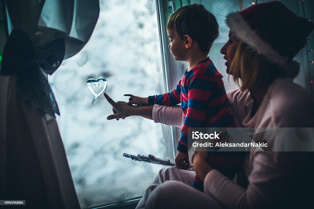 Christmas Eve by the frosted window Photo of mother and her son wearing pyjamas, sitting down next to the frosted window on Christmas Eve and drawing heart shape on the frost Window Stock Photo