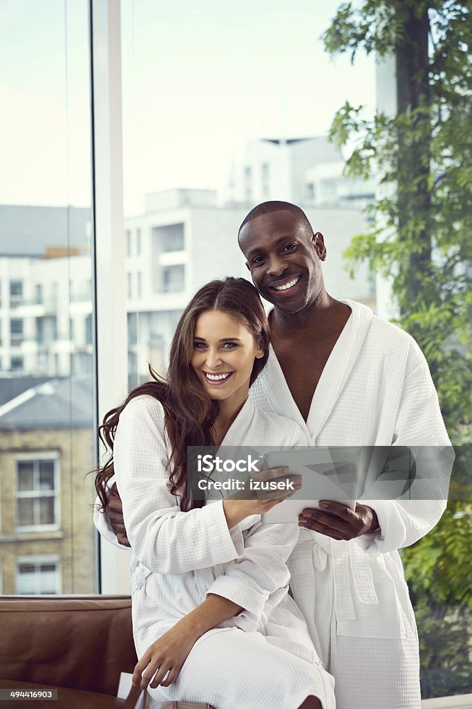 Couple in the morning Friendly afro american man and beautiful brunette wearing white bathrobes standing in their apartment, holding a digital tablet and smiling at the camera. 30-39 Years Stock Photo