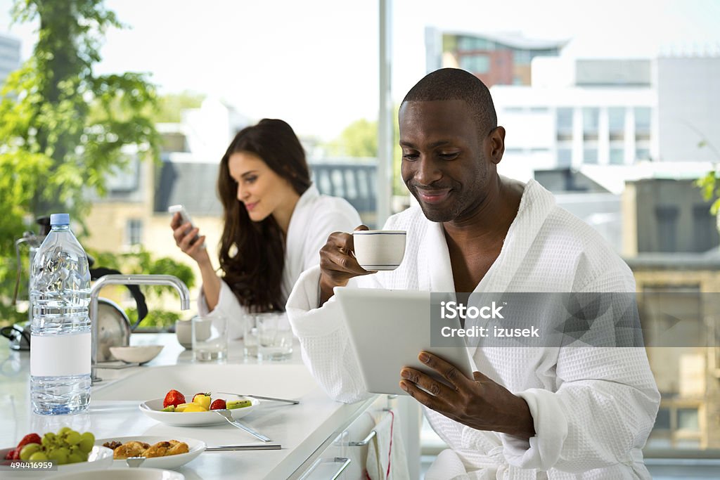 Couple in the morning Afro american man and beautiful brunette wearing white bathrobes having breakfast in their apartment. Focus on the man drinking coffee and using a digital tablet with a woman using smart phone in the background. 30-39 Years Stock Photo