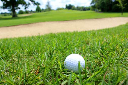 Close-up photo showing a golf ball in the grass on a golf course, with the blades of grass being clearly visible in the shot and stretching into the distance, towards the ever-looming sandy bunker / sand trap hazard.