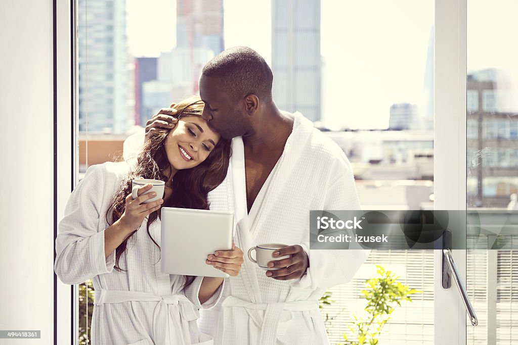 Couple in the morning Afro american man and beautiful brunette wearing white bathrobes standing with cups onf coffee and digital tablet in their aparment in the morning, man kissing a woman. Couple - Relationship Stock Photo