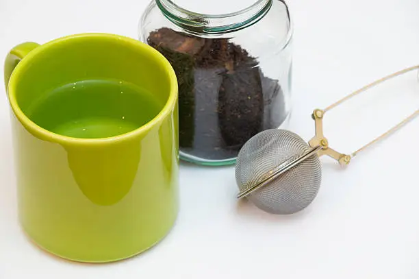 Empty tea infuser and a jar of tea next to each other with a green cup which contains liquid.