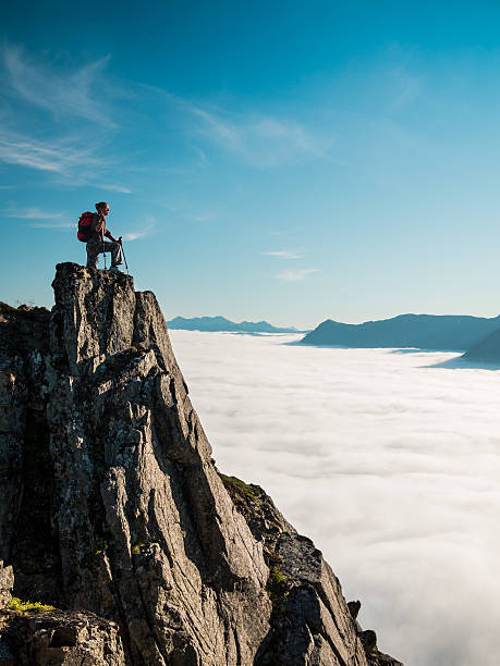 Toned image of an adult woman standing on top mountain Toned image of an adult woman standing on top of a mountain with a backpack and Alpenstocks against mountains in a fog and the blue sky mountain peak stock pictures, royalty-free photos & images