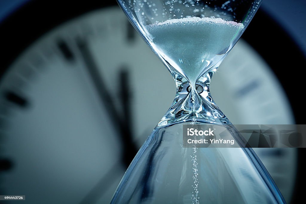 Hourglass Sands of Time Deadline A hourglass with falling sand in front of a clock reaching midnight. Concept photo urgency, and time is running out and deadline is approaching. Close-up of hour glass is photographed in horizontal format with copy space, against a soft-focus clock face in the background. Hourglass Stock Photo