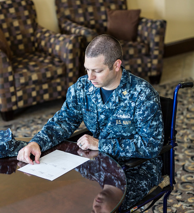 A United States Navy Officer with a physical disability is sitting in a wheelchair in a conference room setting. He is looking down and reading from a document on the table in front of him. The model is wearing a completely authentic U.S. Navy Supply Corps Lieutenant Rank \