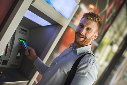 Smiling redhead businessman withdrawing money from a cash machine and looking at the camera.