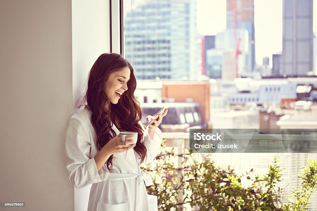 Woman in the morning Beautiful young woman wearing white bathrobe standing with cup of coffee in her apartment in the morning and using a smart phone. High Society Stock Photo