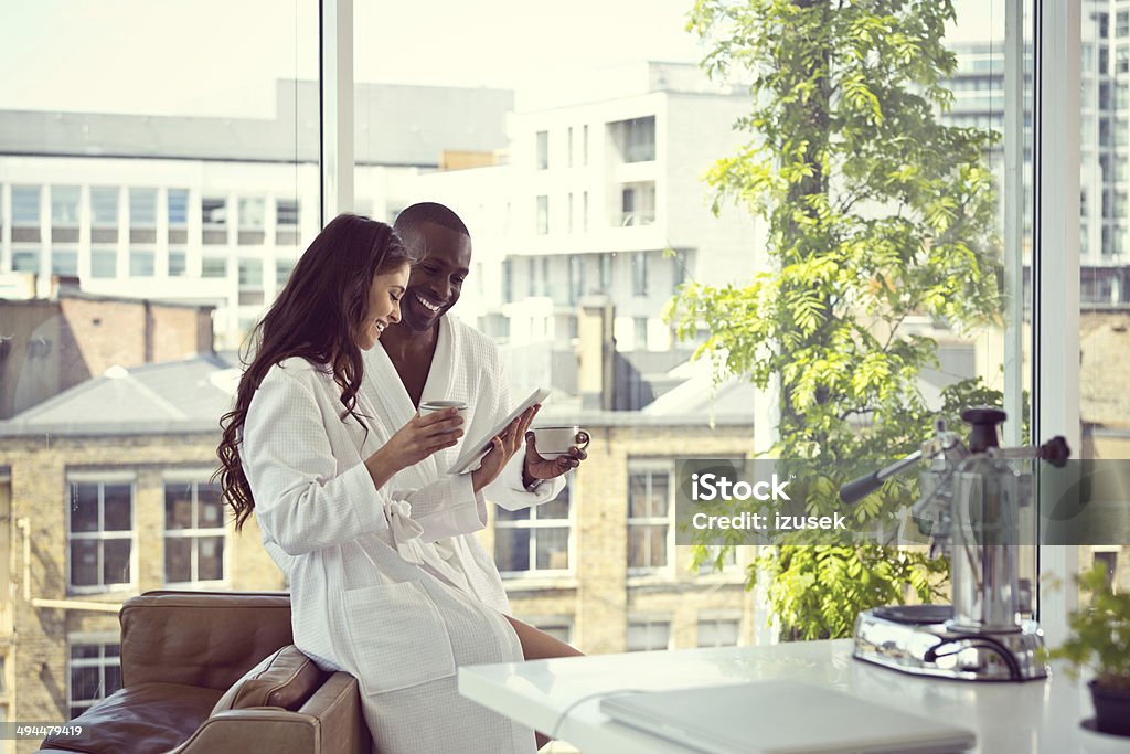 Couple in the morning Afro american man and beautiful brunette wearing white bathrobes drinking coffee and using a digital tablet together in the morning in their apartment. High Society Stock Photo