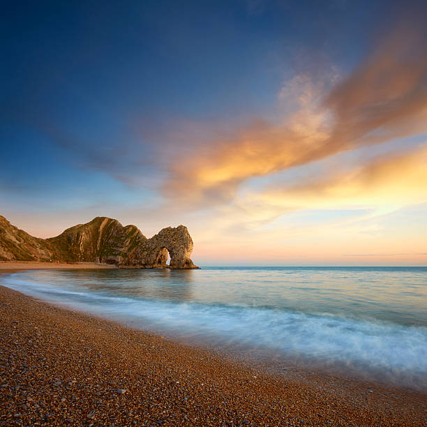 Composite image of Durdle Door on Dorset's Jurassic Coast. A longer exposure captures the movement of the wave whilst overhead a cloud is illuminated by the setting sun. The great limestone arch that makes up Durdle Door can be seen at the end of the beach.