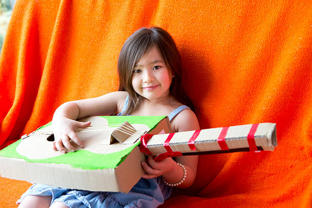 Girl Plays Guitar Chinese girl sits relaxing playing a homemade guitar. She is sitting against an orange throw and smiling at the camera. The guitar is made out of cardboard, tape and string. resourceful stock pictures, royalty-free photos & images
