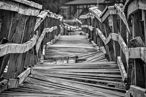 Closeup of a deformed and dilapidated wooden bridge in black and white colors. Broken planks in a very old construction of a vintage bridge over a small river near the village.