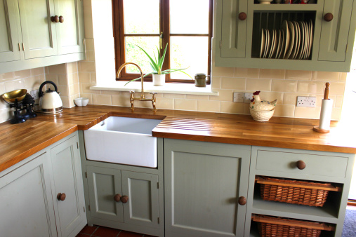 Photo showing a traditional country kitchen, with a large range cooker with gas hob, duck egg green cupboards and wall cabinets, a white ceramic sink (Belfast sink), a terracotta tiled floor, wooden worktops, shelves with storage vegetable baskets, a plate rack, and a pine table and chairs.