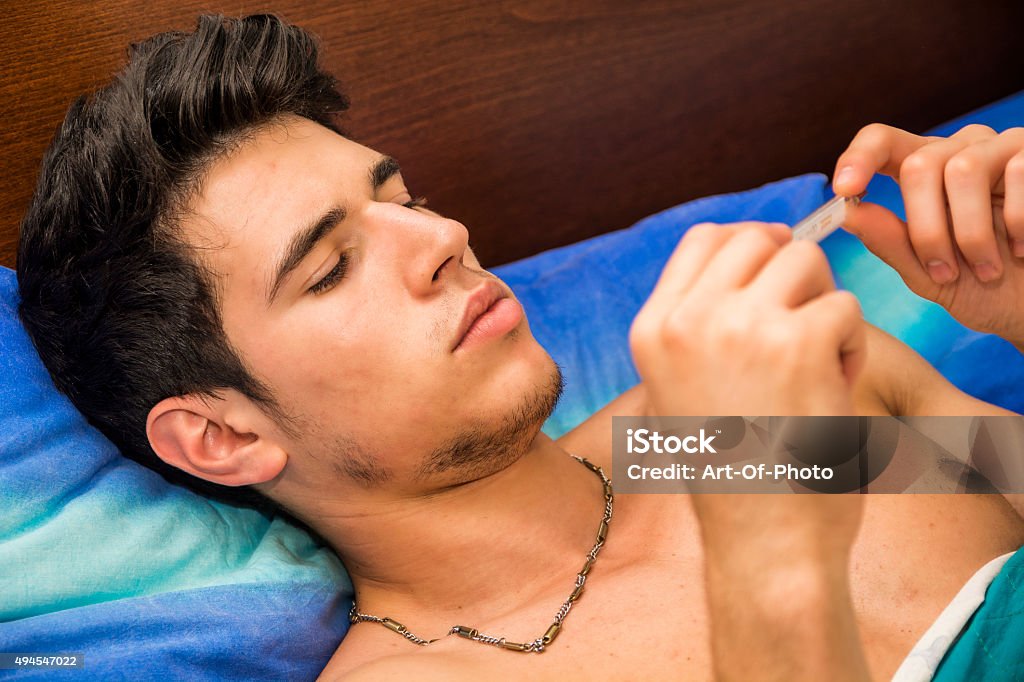 Young man in bed measuring fever with thermometer Young handsome man in bed with a flu or measuring fever with thermometer in his hand, checking the temperature 2015 Stock Photo