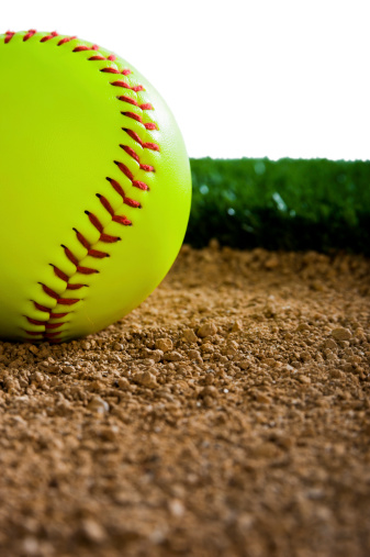 A Close-up of a New Softball in the dirt next to grass against a white background