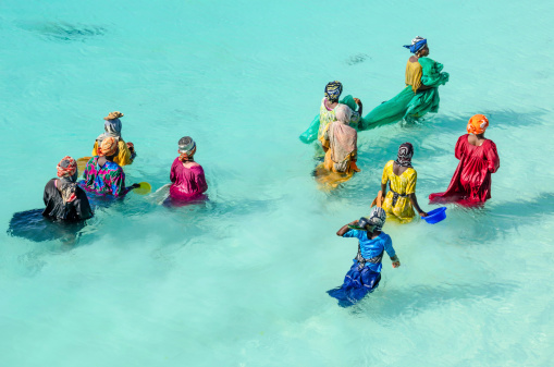 Zanzibar, Tanzania - November 23, 2012: Women with colorful clothes fishing in shallow watter. They are carrying their pots,  with which they hunt small fishes into their net. After a couple of hours they take  home just a full pot of small fishes.