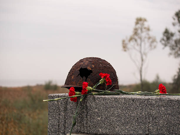 Old rusty military helmet with big hole lies at memorial Old rusty military helmet with a big hole lies at the memorial next to a bouquet of red carnations against the backdrop of an overcast sky and trees patriotic quilt stock pictures, royalty-free photos & images